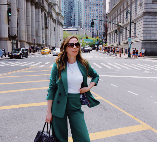 White lady wearing an open teal suite and white shirt with glasses on and a hand bag standing in the street